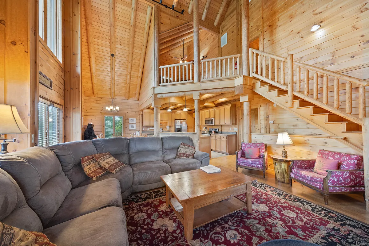 This image showcases a spacious open-plan living area with a large sectional sofa, wooden staircase, high ceilings with exposed beams, and a visible kitchen area complete with colorful armchairs. The room exudes a cozy and welcoming atmosphere, perfect for family gatherings, game nights, and relaxing evenings.