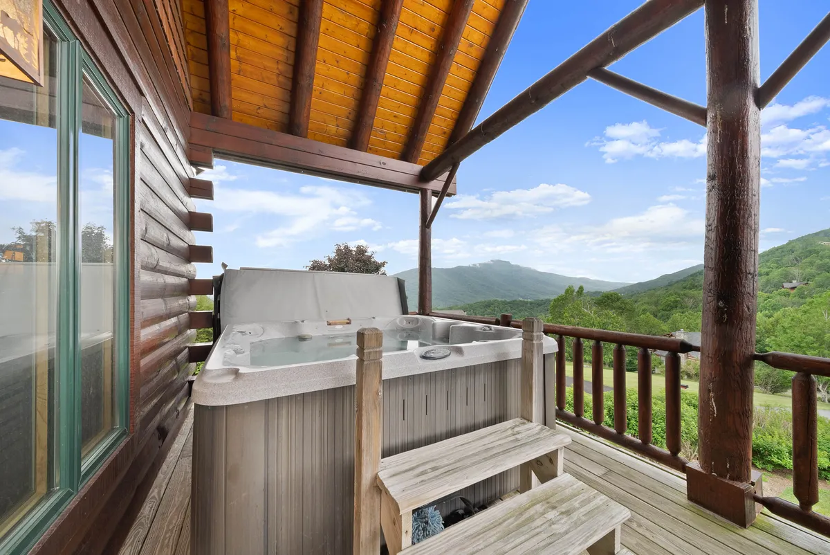 This image features a bubbling hot tub set on a wooden deck, with panoramic mountain views stretching into the distance. The log cabin exterior is visible, and the setting conveys a tranquil, rejuvenating outdoor experience in an elevated alpine environment.