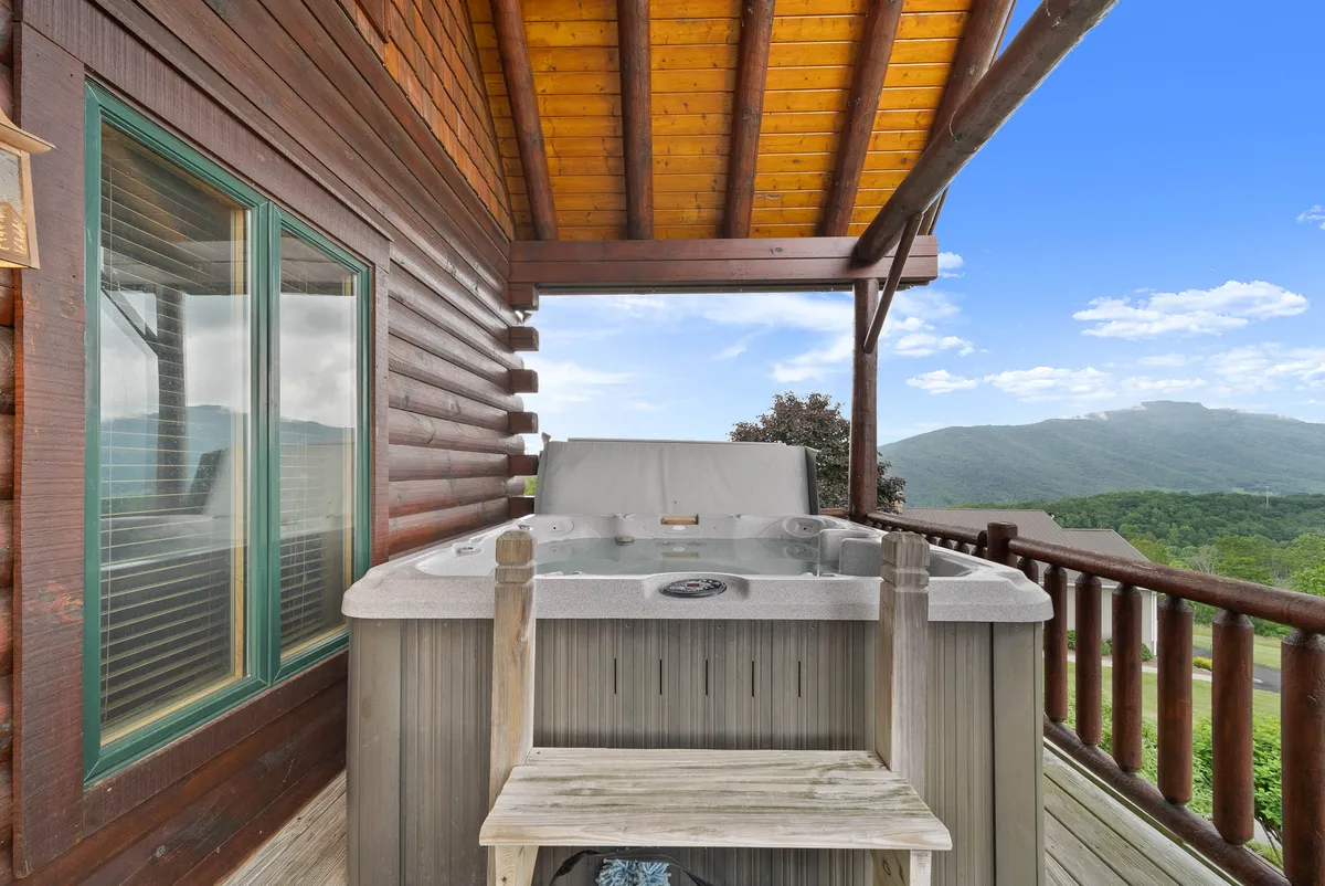 A luxurious hot tub on a wooden deck overlooks sweeping mountain vistas, framed by a classic log cabin structure and sturdy railings. The setting exudes serenity, perfect for unwinding together after a day full of adventures.