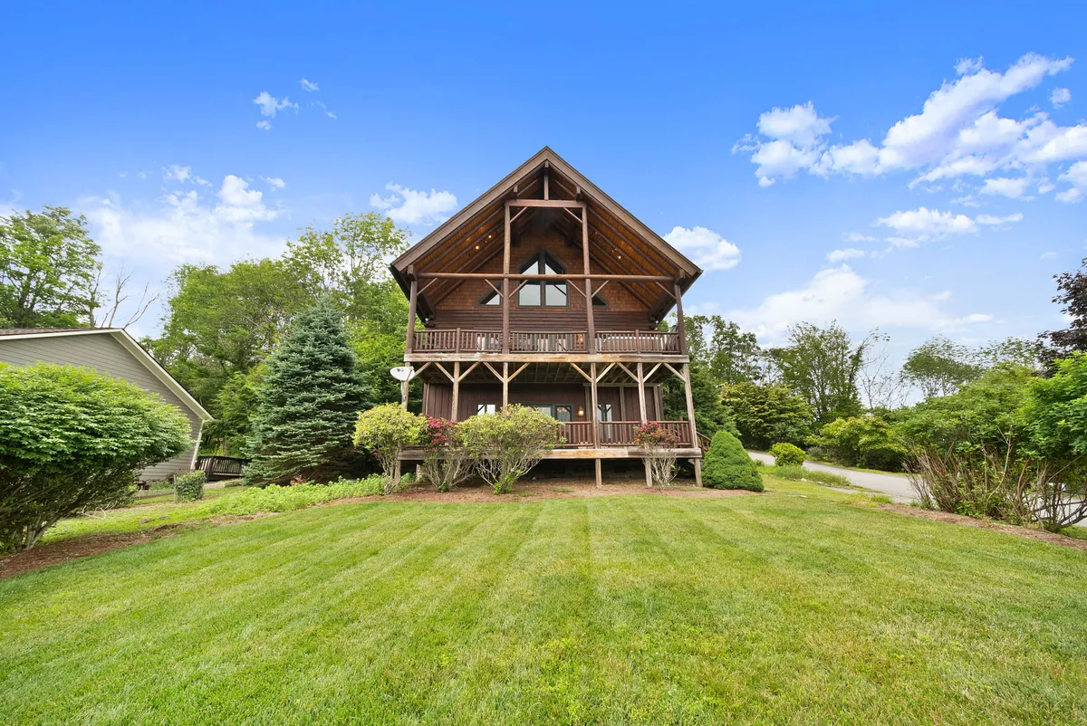 A wide-angle exterior view of the property, showcasing its wooden architecture, expansive green lawn, and inviting front porch. The scene hints at outdoor gatherings, relaxation, and the scenic natural setting that surrounds the home.
