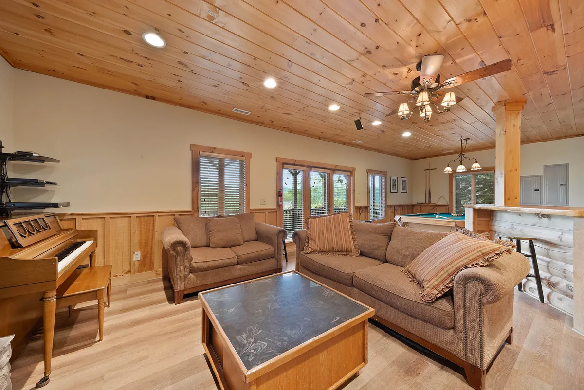 A warm, welcoming room with comfortable sofas, a piano, a pool table, a wooden ceiling, and large windows. The scene suggests lively social gatherings, musical evenings, and friendly pool competitions.