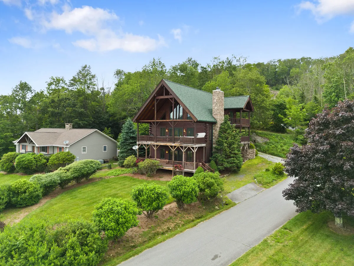 A charming cabin-style home with a green roof sits nestled in lush greenery, its expansive front porch framed by wooden railings. The setting sun casts a golden glow, hinting at peaceful evenings and opportunities for outdoor family gatherings or explorations of nearby nature trails.