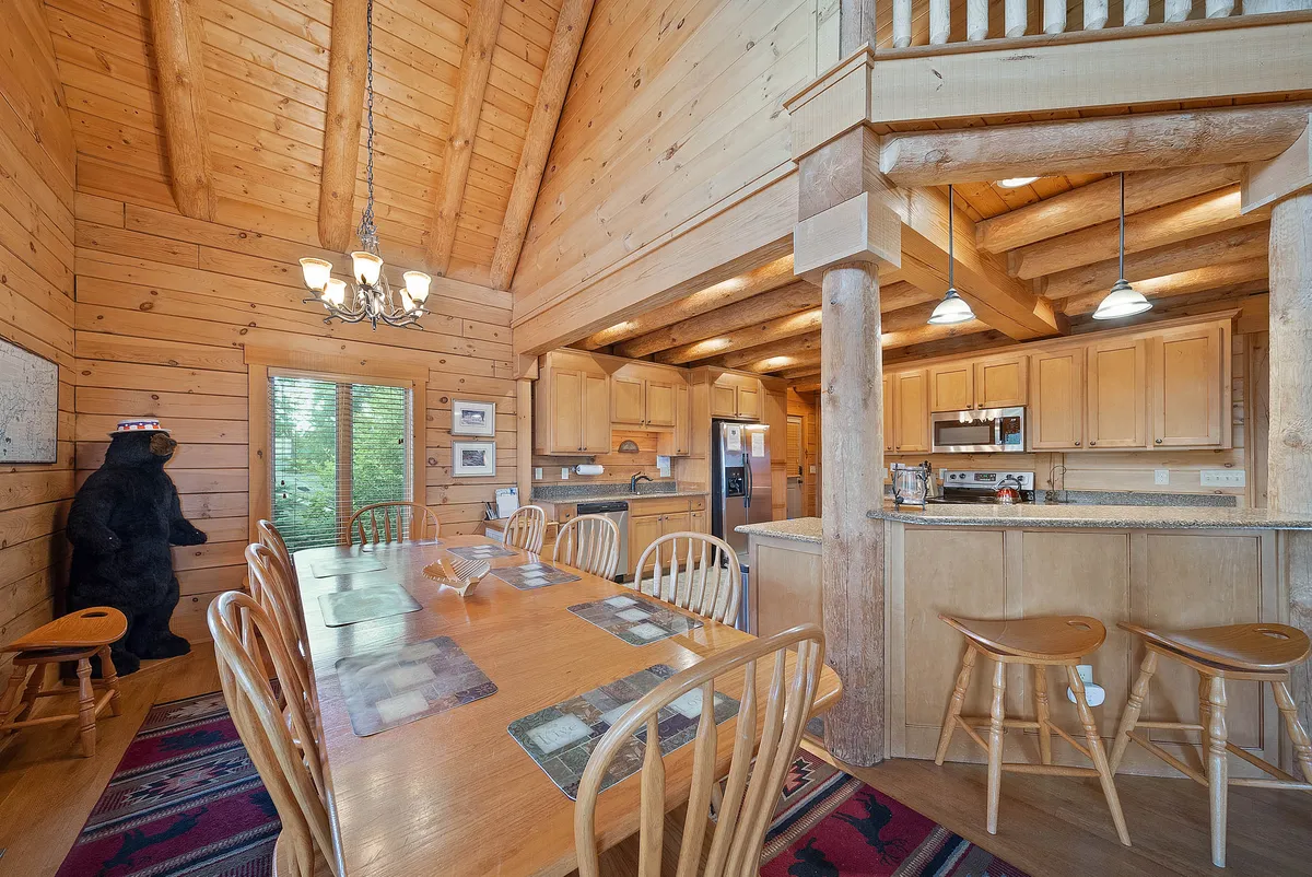 A warm, inviting dining area featuring a large wooden dining table with ample seating, a kitchen in the background, exposed wooden beams, and a whimsical bear statue. The design encourages memorable family meals and entertaining guests.