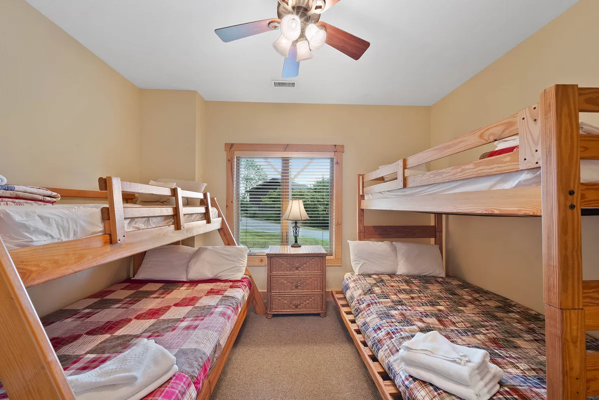 A cozy bunk room with two twin-over-full bunk beds, a bedside table with lamp, and a window filling the room with soft natural light. The space feels playful and perfect for children or family bonding.