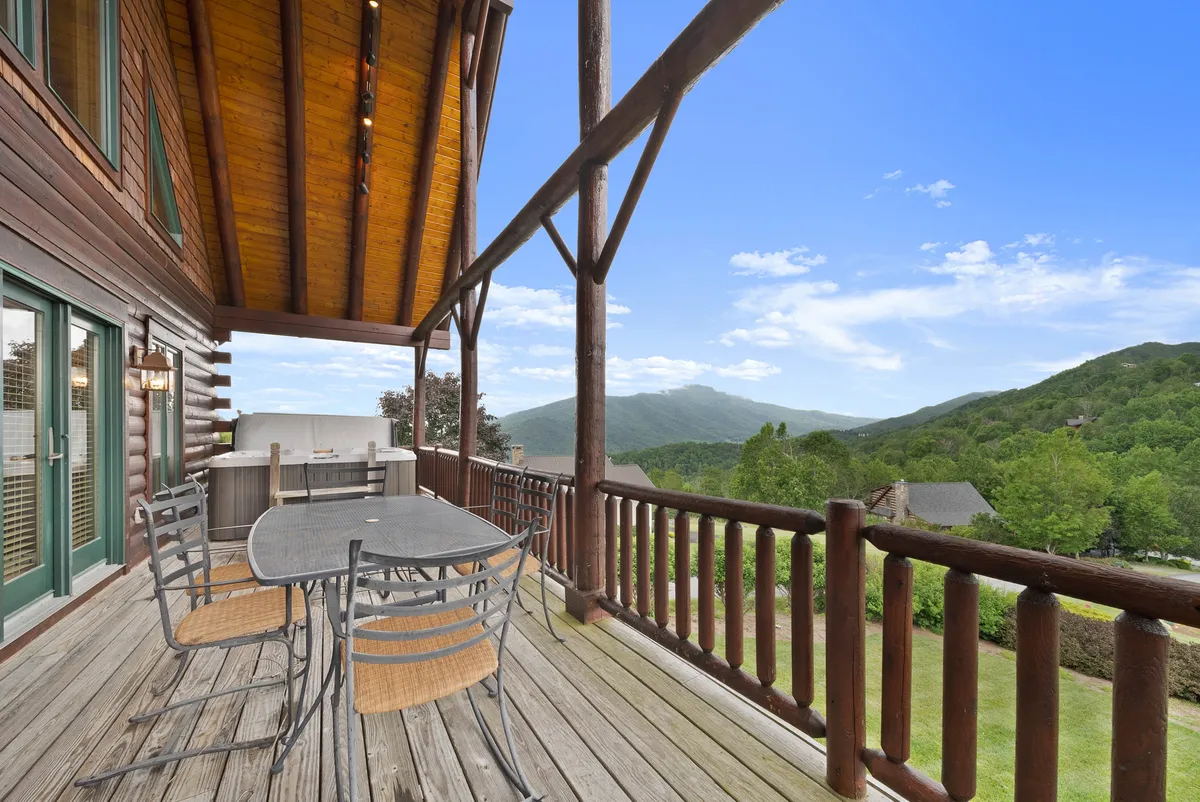 A spacious outdoor dining area on a wooden balcony presents stunning mountain views, with comfortable seating for group meals and a backdrop of peaceful nature. The log cabin’s rustic exterior adds to the inviting ambiance.