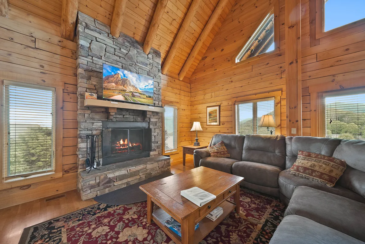 This cozy living room features a striking stone fireplace, large windows that let in natural light, a comfortable couch, a wooden coffee table, and a television displaying a serene nature scene. The arrangement is ideal for relaxing, watching movies, and sharing stories by the fire.